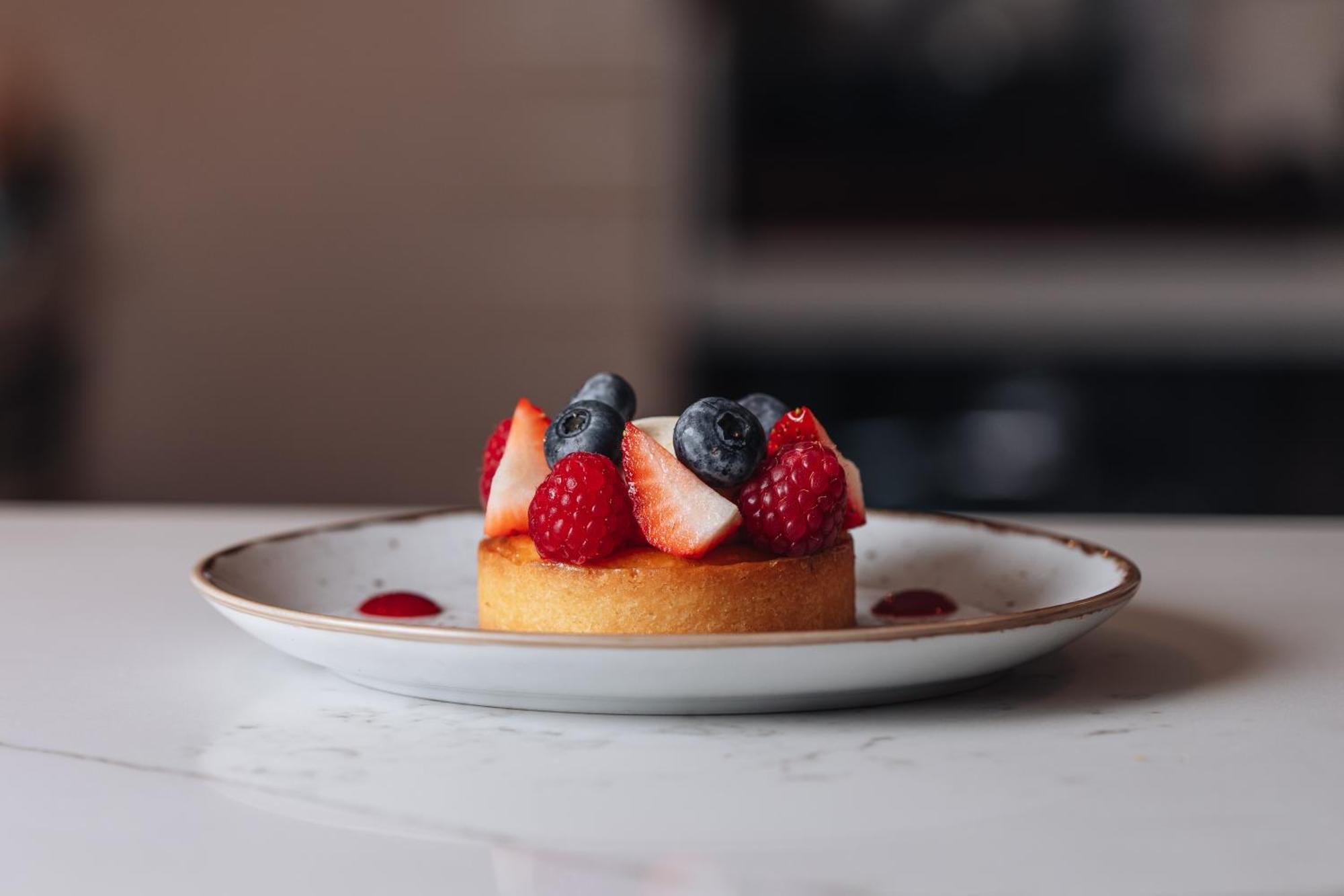 Middle Eight - Covent Garden - Preferred Hotels And Resorts Londra Esterno foto The photo shows a delicious-looking dessert on a white plate. The dessert appears to be a small tart or cake topped with fresh strawberries, raspberries, and blueberries. It has a golden crust, and there are small dollops of a red sauce or fruit pure